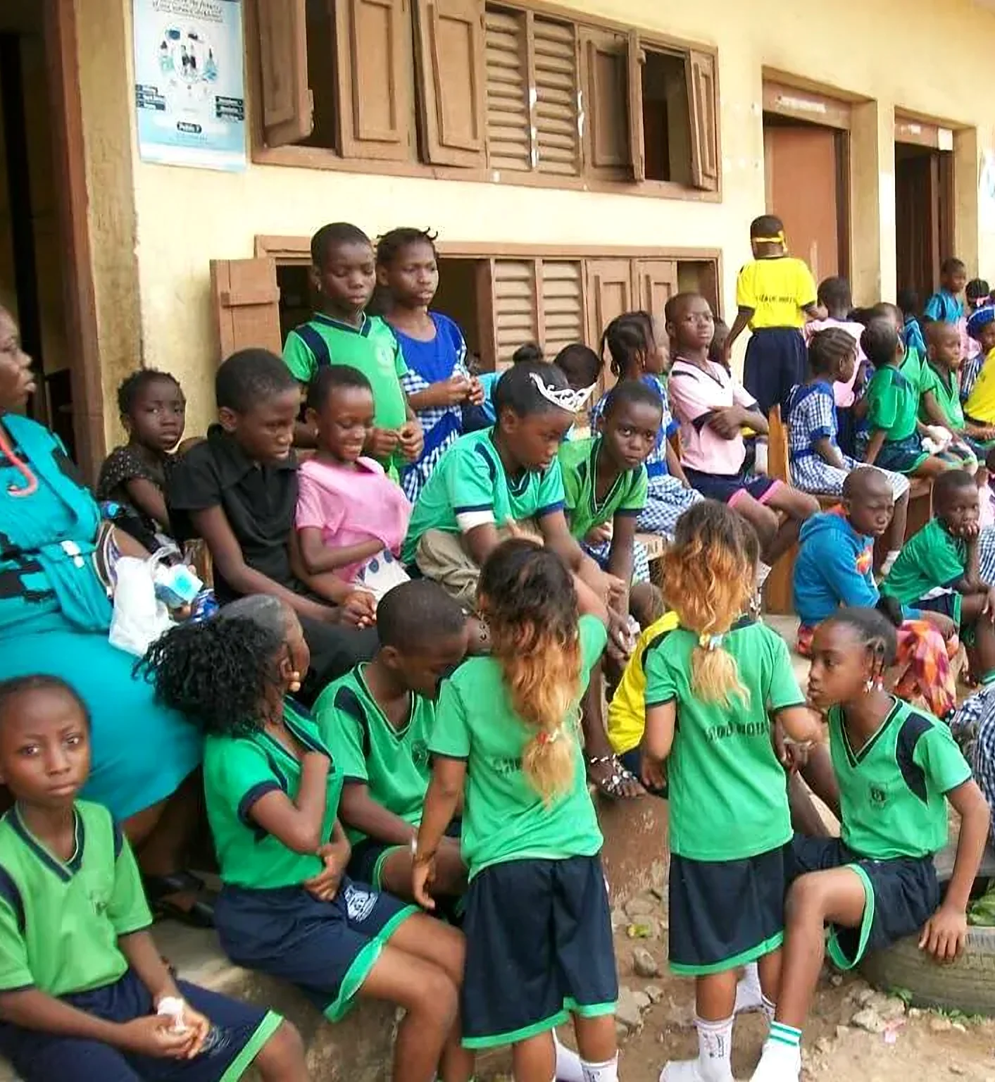 Pupils in green school uniforms sitting together outside their classrooms.